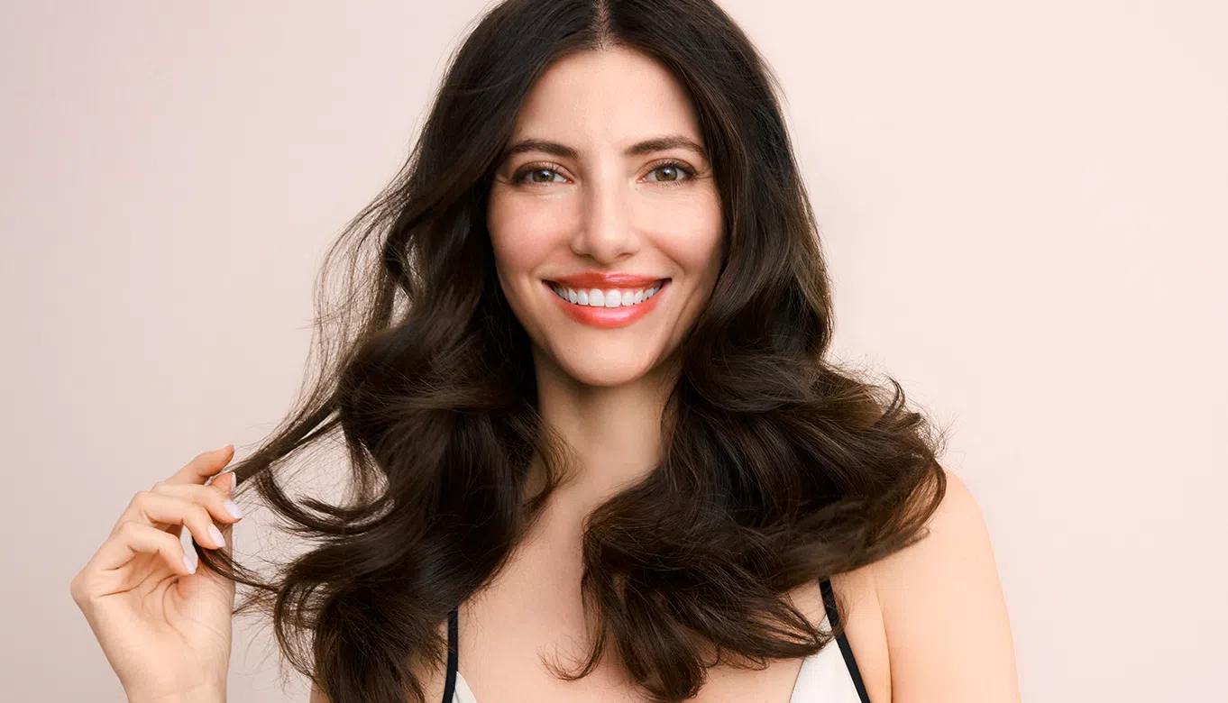 Glimlachende vrouw met lang, donker en volumineus haar houdt een haarlok vast tegen een lichte achtergrond.