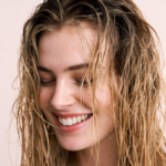 Glimlachende vrouw met nat, net gewassen haar tegen een zachte lichtroze achtergrond.