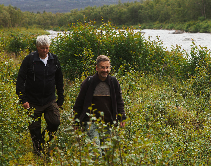 Twee mannen wandelen in de natuur omringd door gras en bloemen.