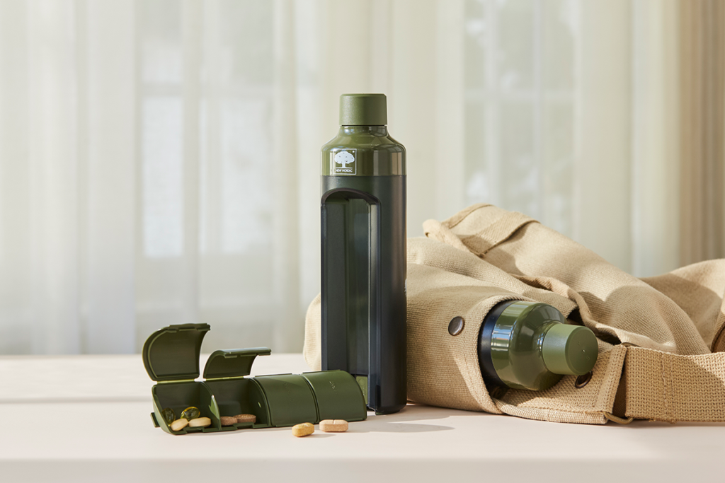 Groene pillenfles met geopende dispenser naast tas en tabletten op een lichte ondergrond.