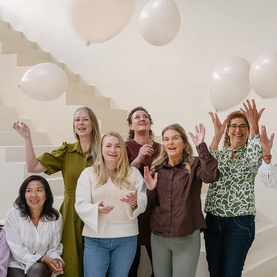 Groep vrouwen gooit witte ballonnen omhoog in een lichte binnenruimte.